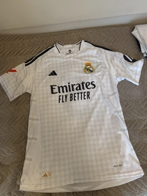 Real Madrid matchtröja - Säljer en vit Real Madrid t-shirt med Bellingham och nummer 5 på ryggen. Tröjan har korta ärmar, svart tryck och klubbmärke på bröstet samt sponsorlogga framtill. Tillverkad i ett lätt och ventilerande material, perfekt för fotboll eller som supporterplagg.