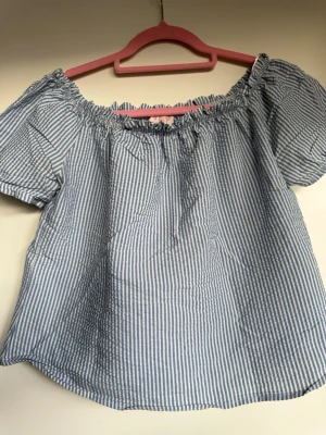 Blårandig offshoulder blus - Säljer en söt blårandig offshoulder blus med korta puffärmar och resår vid halsringningen. Toppen har en luftig passform och är gjord i bomull, perfekt för varma dagar.