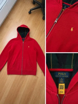 Röd hoodie från Polo Ralph Lauren - Snygg röd hoodie från Polo Ralph Lauren med dragkedja framtill och klassisk gul logga på bröstet. Hoodien har huva med mörkgrönt innerfoder och två fickor fram. Perfekt för en chill och stilren look.