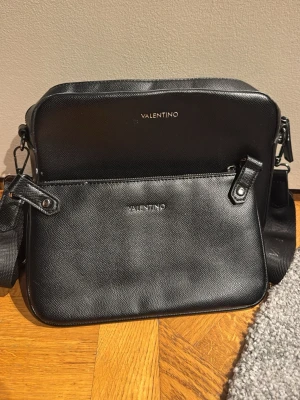 Svart axelväska från Valentino - Snygg svart axelväsk set från Valentino i stilren design. Väskan har justerbar axelrem och är tillverkad i ett slätt, strukturerat konstläder med silverfärgade detaljer. Perfekt storlek för det viktigaste och logga framtill för en exklusiv touch. En liten defekt på lilla väskan, se sista bild!