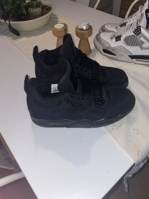 Black Cats Jordan 4 - Säljer ett par helsvarta Air Jordan sneakers med högt skaft och klassisk meshdetalj på sidorna. Skorna har en robust sula och ikoniska detaljer som plastvingar och Jumpman-logga på plösen. Perfekta för dig som gillar streetwear och sportig stil.