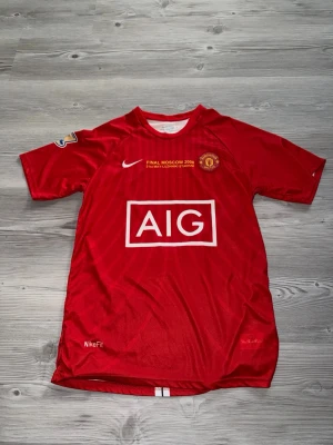 Manchester United  - Röd Manchester United matchtröja från Nike med AIG-logga på bröstet och tryck för Champions League-finalen i Moskva 2008. Tröjan har korta ärmar, klubbmärke och NikeFit-material som andas. Perfekt för fotbollsfans!
