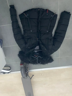 Svart dunjacka med fuskpäls - Snygg svart dunjacka med huva och fuskpäls. Jackan har två dragkedjeförsedda bröstfickor och knäppning framtill. Puffig modell som håller dig varm under kalla dagar. Perfekt för vintern och har en modern, ungdomlig look. 