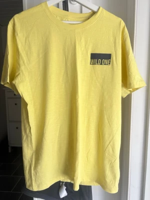 Gul t-shirt med tryck 'Wild One' - Fräsch gul t-shirt med trycket 'WILD ONE' på bröstet. Klassisk rund halsringning och korta ärmar. Tillverkad i mjuk bomull som känns skön mot huden. Perfekt för dig som vill sticka ut med en enkel men cool look.
