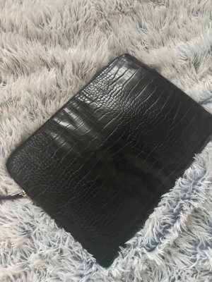 Svart datorfodral i skinnimitation - Snygg svart datorfodral med krokodilpräglat mönster och guldfärgad dragkedja. Väskan har en stilren och modern look, perfekt att bära i handen eller under armen. Materialet är glansigt och ger en lyxig känsla.