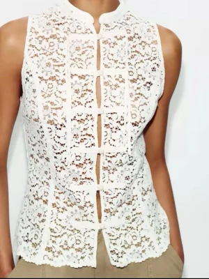 Vit spetsblus med knappar från zara - Supersnygg vit blus i spets med dekorativa knappar framtill och hög krage. Ärmlös modell med vackert blommigt mönster och figurnära passform. Perfekt för dig som vill ha en romantisk och trendig topp till sommaren.