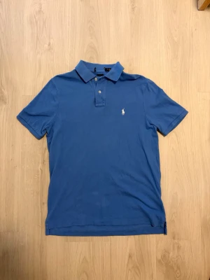 Ralph Lauren Pike - Klassisk blå pikétröja med korta ärmar och krage från Polo Ralph Lauren. Tröjan har två knappar vid halsen och en liten broderad logga på bröstet. Tillverkad i mjuk bomull som känns skön mot huden. Perfekt till jeans eller shorts. Etiketten i nacken (Se bild) har gått av, inget som syns när man bär piken.