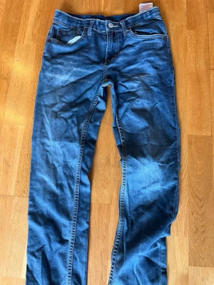 Blå Levi jeans - Säljer ett par klassiska blå jeans med raka ben och snygga slitningar framtill. Jeansen har fem fickor, kontrastsömmar och en patch bak på linningen. Materialet är denim i bomull och passformen är avslappnad men ändå stilren.