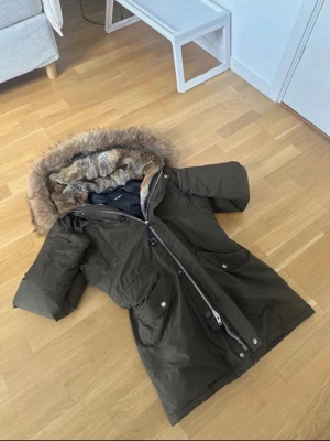 Olivgrön parkas med fuskpäls Hawk lux - Snygg olivgrön parkas med stor huva fodrad med mjuk fuskpäls i bruna och grå toner. Jackan har dragkedja och knappar framtill, stora fickor och en längre passform som håller dig varm under kalla dagar. Perfekt för dig som vill ha både stil och funktion. Äkta päls av både kanin och räv Aldrig anvand och är helt ny Köpte för 5000 kr så säljer därför för minst 3500 Säljer för jag redan köpt ny vinterjacka