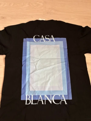 Svart t-shirt från Casablanca - Snygg svart t-shirt från Casablanca med stor grafisk print i blå toner och vit text på ryggen samt mindre tryck på bröstet. Klassisk rund hals och korta ärmar. Perfekt för dig som gillar stilrena men ändå unika plagg.