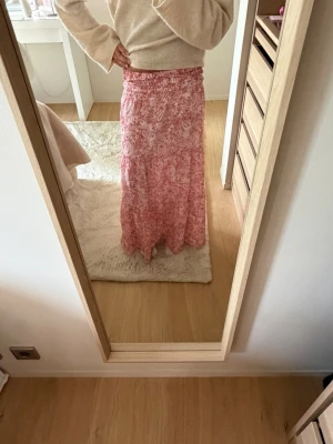 Rosa långkjol med blommigt mönster - Säljer en rosa långkjol med vitt blommigt mönster. Kjolen har en avslappnad passform och resår i midjan för extra komfort. Perfekt för vår och sommar, och materialet känns lätt och luftigt.🩷passar mig som brukar ha xs, längden är bra om man är runt 155-165 beroende hur man villa ty den ska vara i längden!💕