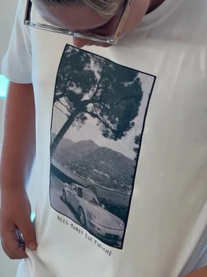 Vit t-shirt med Porsche-tryck - Säljer en vit t-shirt med ett stort grafiskt tryck på bröstet som föreställer en Porsche och texten 'Need money for Porsche'. T-shirten har klassisk passform och rund hals. Perfekt för dig som gillar bilar och streetstyle.