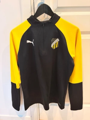 Svart och gul träningströja  och byxor Puma Häcken - Snygg långärmad träningströja från Puma med BK Häcken-logga på bröstet. Tröjan är svart med gula ärmar och har en halv dragkedja framtill. Tillverkad i funktionsmaterial som passar perfekt till fotbollsträning eller annan sport. Som också kommer med passande byxor vilket gör det till en hel traicksuit 