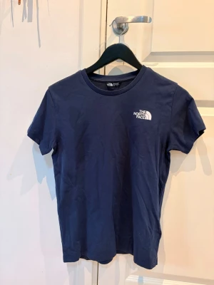 Marinblå t-shirt från The North Face - Snygg marinblå t-shirt från The North Face med klassisk logga tryckt i vitt på bröstet. T-shirten har rund halsringning och korta ärmar. Tillverkad i mjuk bomull som känns skön mot huden. Perfekt till jeans eller shorts.