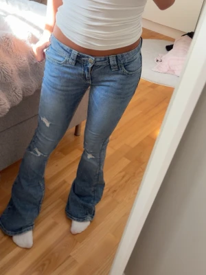 Blå bootcut jeans med slitningar - Säljer ett par ljusblå bootcut jeans med slitna detaljer på benen och broderade bakfickor. Jeansen har låg midja och kommer ifrån gina tricot 14+!💕 Eftertraktade jeans!