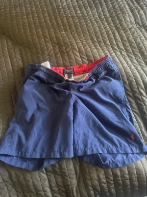 Blå badbyxor från Polo Ralph Lauren - Snygga blå badbyxor från Polo Ralph Lauren med röd insida och klassisk logga broderad i rött på benet. Elastisk midja och avslappnad passform, perfekta för stranden eller poolen.