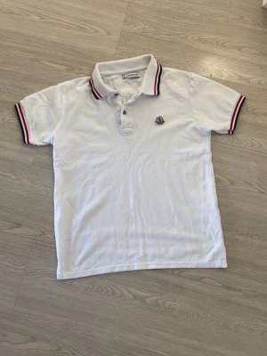 Vit pikétröja med rödblå detaljer - Klassisk vit pikétröja från Moncler med korta ärmar och krage. Tröjan har rödblå ränder på krage och ärmslut samt Moncler-logga på bröstet. Perfekt till en avslappnad men stilren look.
