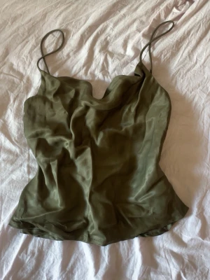 Olivgrön linnetopp med knappar - Snygg olivgrön satin linnetopp med tunna axelband och dekorativa knappar på sidan. Toppen har en mjuk, något lös passform och ett lätt glansigt material som ger en lyxig känsla. Perfekt att styla med jeans eller kjol för en trendig look.