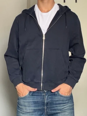 Mörkblå zip hoodie från Weekday - Säljer en mörkblå hoodie från Weekday med dragkedja framtill, huva med dragsko och två stora fickor. Tillverkad i mjukt bomullsmaterial och har ribbade muddar vid ärmslut och nederkant. Perfekt för en avslappnad och stilren look.