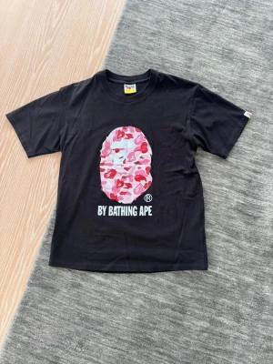 Svart BAPE t-shirt med camo-print - Säljer en svart t-shirt från BAPE med det ikoniska ap-huvudet i rosa, röd och vit camouflage på bröstet. Under trycket står det 'BY BATHING APE'. Klassisk passform och rund hals. Tillverkad i Japan.