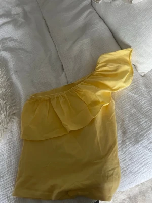 Gul oneshoulder topp med volang - Snygg gul oneshoulder topp med volangdetalj längs axeln. Perfekt för dig som vill sticka ut med en färgglad och trendig look. Tillverkad i mjukt bomullsmaterial som känns skönt mot huden.