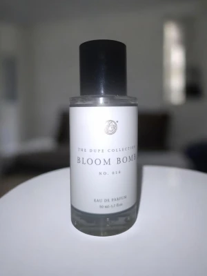 Bloom Bomb Eau de Parfum 45 ml - Fräsch parfym från The Dupe Collection, Bloom Bomb No. 014. Flaskan är genomskinlig med svart lock och stilren vit etikett. Innehåller ca 45 ml Eau de Parfum. Perfekt att ha i väskan eller på sminkbordet. Snygg och modern design.