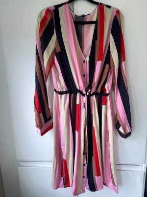 Randig långärmad klänning med knappar - Färgglad klänning med vertikala ränder i rosa, rött, beige och marinblått. Klänningen har v-ringning, knappar framtill, lång ärm och ett knytband i midjan för en snygg passform. Tillverkad i ett mjukt, lätt material. 