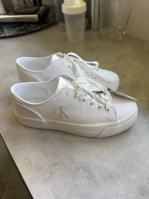 Vita sneakers från Calvin Klein - Stilrena vita sneakers från Calvin Klein med lågt skaft och klassisk snörning. Skorna har en tjock vit sula och är tillverkade i canvasmaterial. Diskret cK-logga på sidan och bak på hälen för en clean look. Helt nya, nypris 899 nu 750. Storlek 40