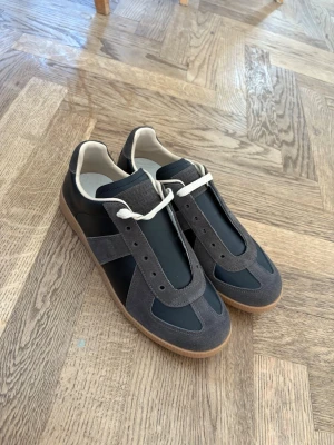 Maskin margiela black/brown - Säljer ett par stilrena sneakers i svart med grå mockadetaljer och vita snören. Skorna har en klassisk siluett med gummisula och passar perfekt till streetwear eller casual outfits. Materialmixen av mocka och syntet ger en schysst kontrast.
