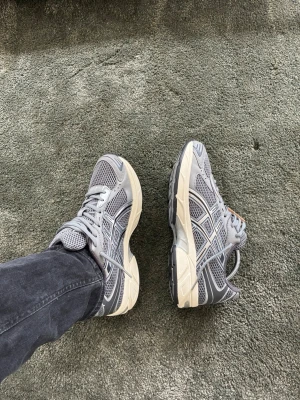 Grå sneakers från Asics - Snygga grå sneakers från Asics med mesh- och syntetpaneler för bra andningsförmåga. Skorna har vita och mörkgrå detaljer samt en beige sula som ger en retrovibe. Klassisk snörning och låg profil gör dem perfekta till streetwear och casual outfits. Kan nog också passa 44
