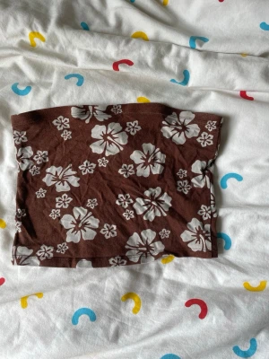 Brun blommig bandeau tubtopp - Säljer en brun bandeau tubtopp med vita blommor i ett somrigt mönster. Toppen är ärmlös och har en enkel, rak passform. Perfekt för varma dagar och enkel att styla med jeans eller kjol.