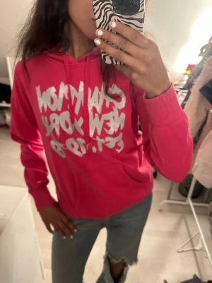 Rosa hoodie med tryck och huva - Säljer en snygg rosa hoodie med vit text på bröstet. Hoodien har en klassisk huva med dragsnören och en stor magficka framtill. Perfekt för dig som gillar streetwear och vill ha en färgklick i garderoben. Passar till jeans eller mjukis.