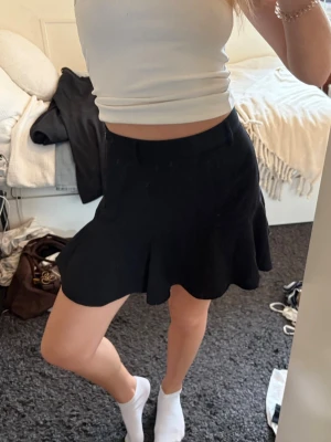 Svart kortkjol med volangkant - Säljer en svart kortkjol med volangkant och lätt utsvängd passform. Kjolen har en enkel midja utan synliga detaljer och är perfekt för dig som gillar en stilren men ändå lekfull look. Materialet känns mjukt och följsamt.