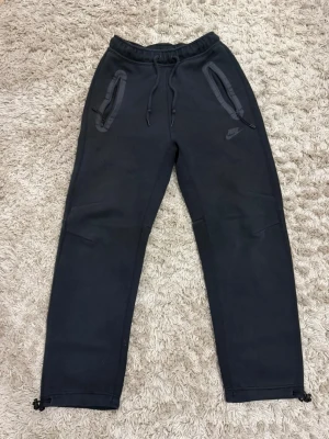 Svarta mjukisbyxor från Nike - Svarta mjukisbyxor från Nike med dragsko i midjan och dragkedjeförsedda fickor. Byxorna har raka ben och justerbara benslut. Tillverkade i mjukt material som passar perfekt för chill eller träning. Diskret Nike-logga på ena benet.