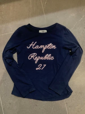 Mörkblå långärmad topp Hampton Republic - Mörkblå långärmad topp från Hampton Republic med texten 'Hampton Republic 27' i glittriga bokstäver på bröstet. Klassisk rund halsringning och mjukt material som känns skönt mot huden. Perfekt till jeans eller kjol för en avslappnad look.