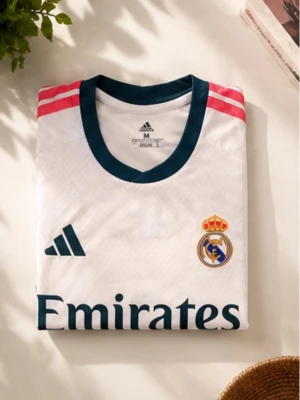 Real Madrid matchtröja vit - Säljer en vit Real Madrid matchtröja från Adidas med mörkgröna detaljer och rosa ränder på axlarna. Tröjan har klubbmärke, Adidas-logga och Emirates Fly Better-tryck på bröstet. Tillverkad i lätt och ventilerande material, perfekt för fotboll eller träning.