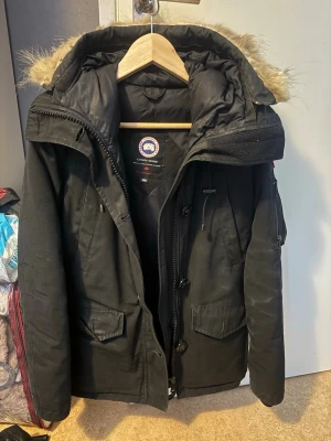 Svart parkas från Canada Goose - Säljer en svart parkas från Canada Goose med stor huva och avtagbar fuskpäls. Jackan har dragkedja, knappar, flera fickor och är fodrad för extra värme. Perfekt för kalla dagar och riktigt snygg till vintern.