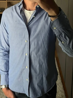 J.Lindeberg skjorta i blå/vitrandig - stl M - Skjorta från J.Lindeberg i 100% bomull. Smal passform med bred krage. Tunt tyg, funkar till mycket olika plagg. Personen på bilden är 189cm, väger 78kg.  Använd men i mycket fint skick, inga hål eller fläckar.  Rök- och djurfritt hem. Skriv gärna om du vill ha fler bilder eller undrar något!