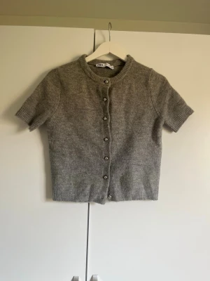 Grå kortärmad kofta från Zara - Mysig grå kofta från Zara med korta ärmar och rund hals. Koftan har ribbade kanter och knäpps med silvriga knappar framtill. Perfekt att slänga över en topp för en enkel men stilren look.