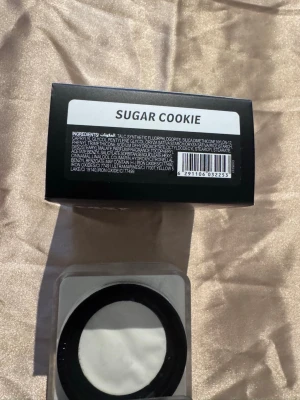 Huda Beauty Easy Bake Sugar Cookie - Translucent löspuder i nyansen Sugar Cookie från Huda Beauty. Kommer i en fyrkantig burk med svart lock och rosa text. Perfekt för att fixera foundation och ge en matt finish. Pudret har en ljus, nästan vit ton och känns lätt på huden.