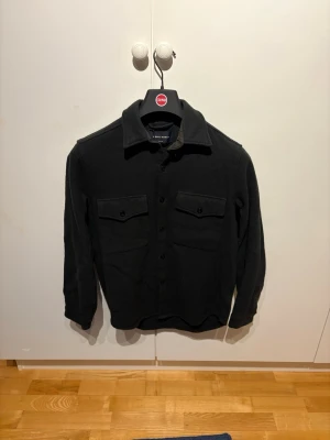 Svart overshirt från A Day's March - Stilren svart overshirt från A Day's March i ullmix. Klassisk krage, knäppning framtill och två bröstfickor med lock och knapp. Perfekt lager-på-lager-plagg för en clean och avslappnad look.