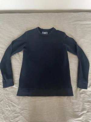 Mörkblå sweatshirt med rund hals - En stilren mörkblå sweatshirt med rund halsringning och långa ärmar. Tröjan har ribbade muddar vid ärmslut och nederkant, vilket ger en klassisk och avslappnad look. Perfekt att matcha med jeans eller joggers för en enkel och snygg outfit.