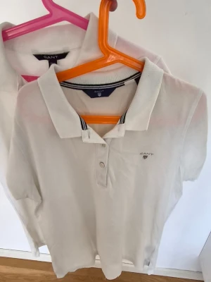 Vit pikétröja från Gant, storlek S/xs - Klassisk vit pikétröja från Gant i storlek xs/S. Tröjan har krage med smala svarta och vita ränder, knappar framtill och diskret Gant-logga broderad på bröstet. Perfekt till jeans eller shorts för en clean look. 