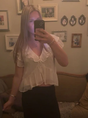 Söt Nelly blus - Söt vit blus med volangdetaljer längs urringningen och ärmarna. Toppen har korta ärmar, v-ringning. Den är lätt och luftig, perfekt för varma dagar och ger en romantisk vibe.
