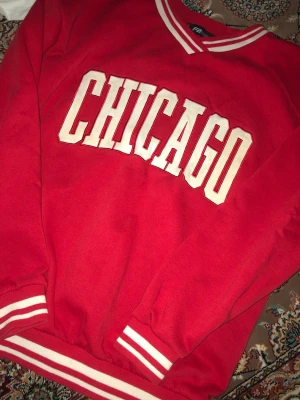 Röd sweatshirt med CHICAGO-tryck - Riktigt snygg röd sweatshirt med stort vitt CHICAGO-tryck på bröstet. Tröjan har vita ränder på krage, ärmslut och nederkant för en sportig look. Perfekt för dig som gillar streetwear och college-stil.