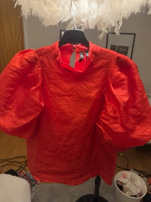 Röd puffärmad blus från PIECES - Trendig röd blus från PIECES med stora puffärmar och hög hals. Tillverkad i ett lätt, strukturerat tyg som ger en cool look. Knäppning i nacken med knappdetalj. Perfekt för dig som vill sticka ut med färg och volym.