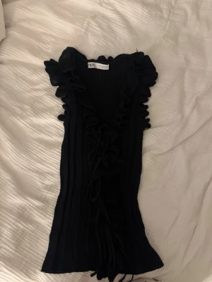 Svart ribbad topp med volang från Zara - Snygg svart ribbad topp från Zara med volangdetaljer längs ärmhål och framkant. Toppen är ärmlös och har en figurnära passform med dekorativ snörning framtill. Perfekt för dig som gillar trendiga detaljer och en chic look.