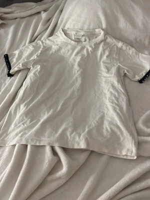 Vit t-shirt med pärldetaljer Zara - Säljer en vit t-shirt från Zara med korta ärmar och svarta pärldetaljer vid ärmsluten. T-shirten har en klassisk passform och är gjord i mjuk bomull, perfekt för en clean och stilren look. 