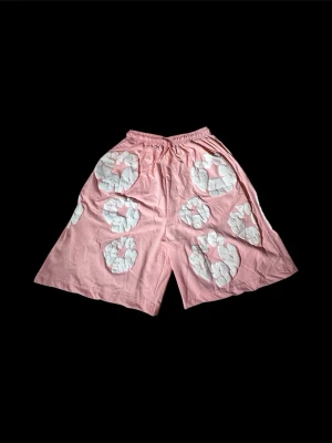 Denim Tears Shorts – Rosa/Vita – Storlek M - De har den ikoniska bomullsblomsdesignen i vitt mot en rosa bas, vilket gör dem väldigt unika och lätta att känna igen. Perfekta för sommaren eller för dig som gillar streetwear med en stilren men iögonfallande look.                                         Shortsen har en bekväm passform med elastisk midja och passar både till en enkel t-shirt och en hoodie. Materialet känns skönt och de är både snygga och bekväma att använda.