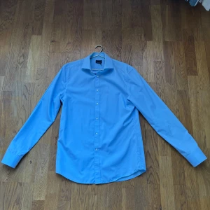 Blå skjorta med långa ärmar - Snygg blå skjorta med långa ärmar och klassisk krage. Perfekt för en stilren look. Skjortan har knappar framtill och är tillverkad i ett lätt och bekvämt material.